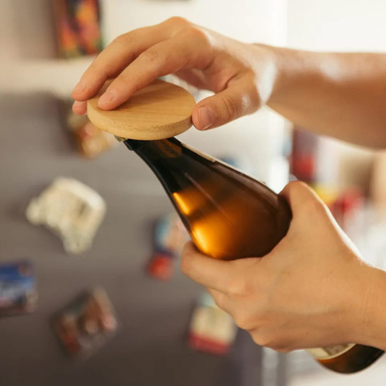 A drink bottle opener made of bamboo, featuring a circular design and held by a hand, with a dark bottle in view.