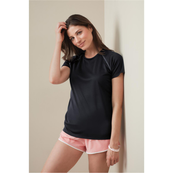 A woman in a black active t-shirt poses against a wall, wearing pink shorts. The shirt features short sleeves and has a logo.