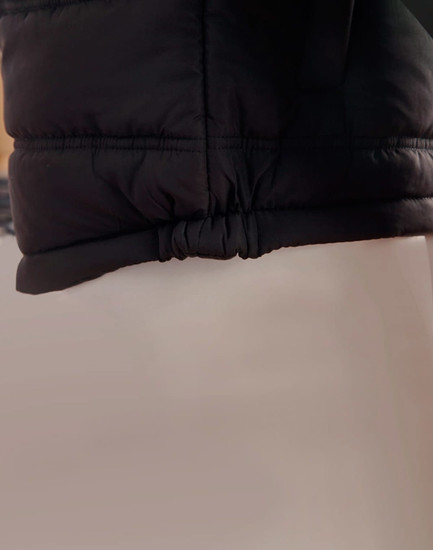 A black heavy quilted puffer jacket featuring a cinched hem and a smooth fabric texture. It has a logo visible.