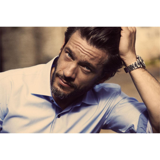 A stylish men's button-up shirt in light blue, featuring a logo, worn by a man with a beard.