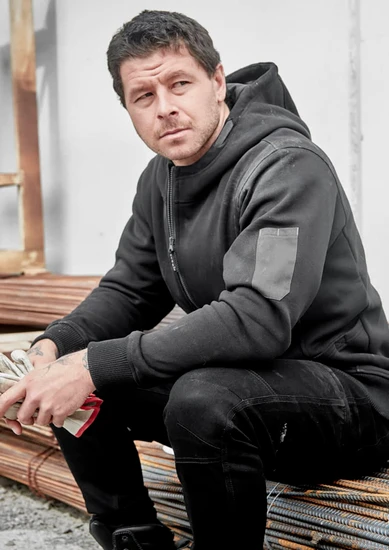 A black unisex zip front hoodie with multi-pockets, featuring a logo, worn by a seated man.