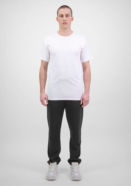 A man stands wearing a plain white t-shirt and black track pants. The track pants feature a logo.