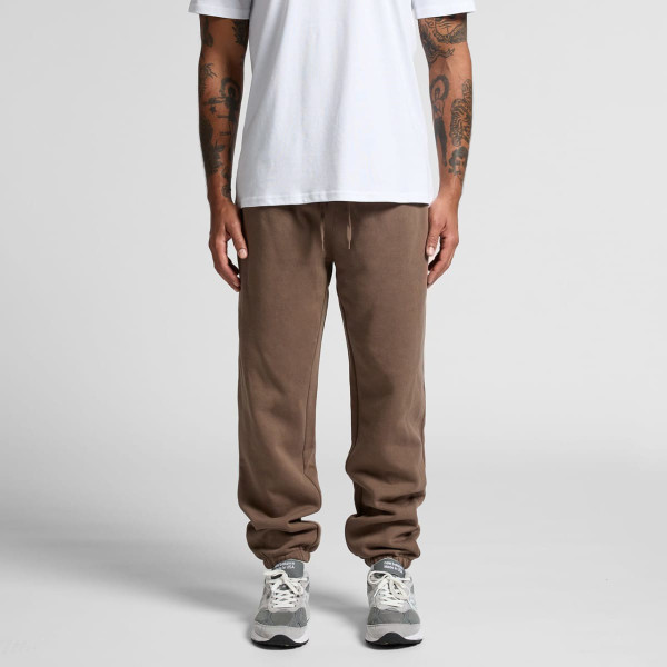 Brown track pants with a relaxed fit and an elastic waistband, paired with grey sneakers. The model wears a plain white t-shirt.