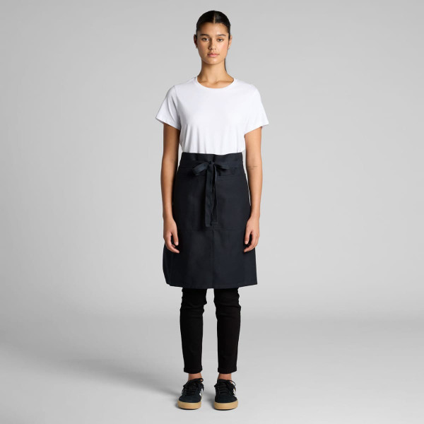 A half apron in black, featuring a front pocket and a tied waistband, displayed on a model wearing casual attire.