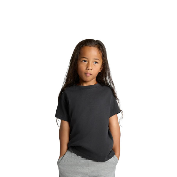 A black kids' heavy faded tee with short sleeves, worn by a child with long hair, standing with hands in pockets.