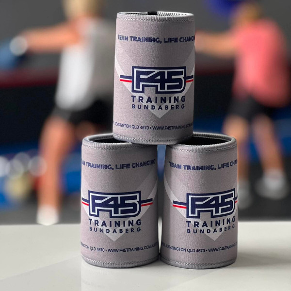 Three grey neoprene can coolers stacked in a pyramid on a white surface, with a blurred gym scene of people exercising in the background.