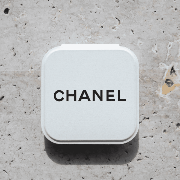 A compact mirror in a white case with a logo on the top, set against a textured stone background.