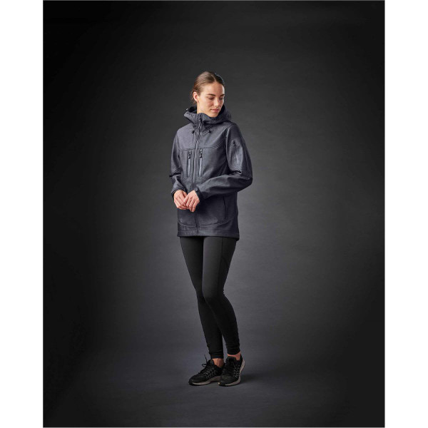 A woman wearing a dark grey softshell jacket, paired with black leggings and black shoes, on a dark background.