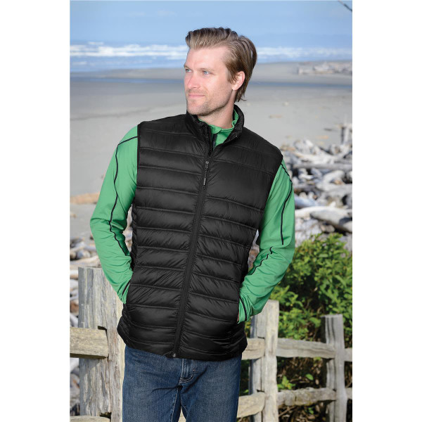 A man wearing a black thermal vest over a green long-sleeve shirt, standing by a beach with driftwood in the background.