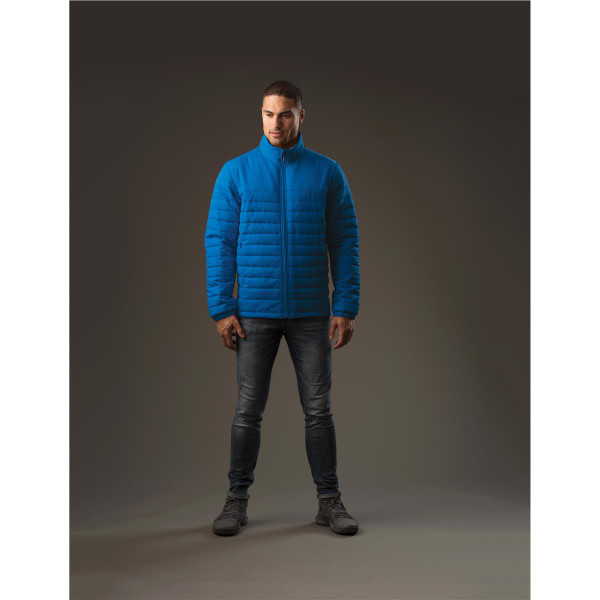 A man stands wearing a blue quilted jacket, paired with jeans and boots, with a neutral background.