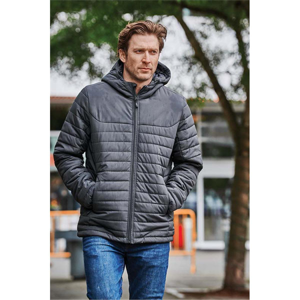 A man wearing a dark grey quilted hoody, walking outdoors surrounded by greenery. The hoody features a logo.
