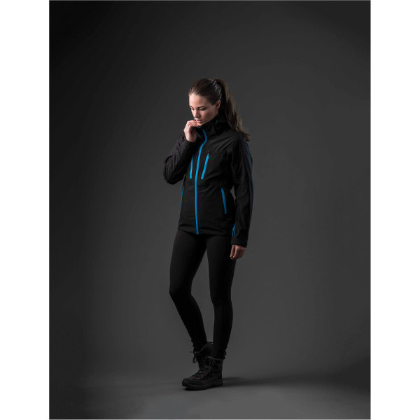 A woman wearing a black jacket with blue accents, styled with black leggings and boots, stands against a dark background.