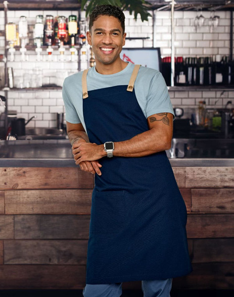 A woman stands against a brick wall, wearing a beige Brunswick Bib Apron with black straps. The apron features a pocket.