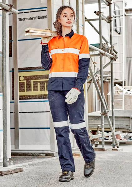 A woman in an orange and navy work shirt and matching pants stands on a construction site, holding wooden planks.