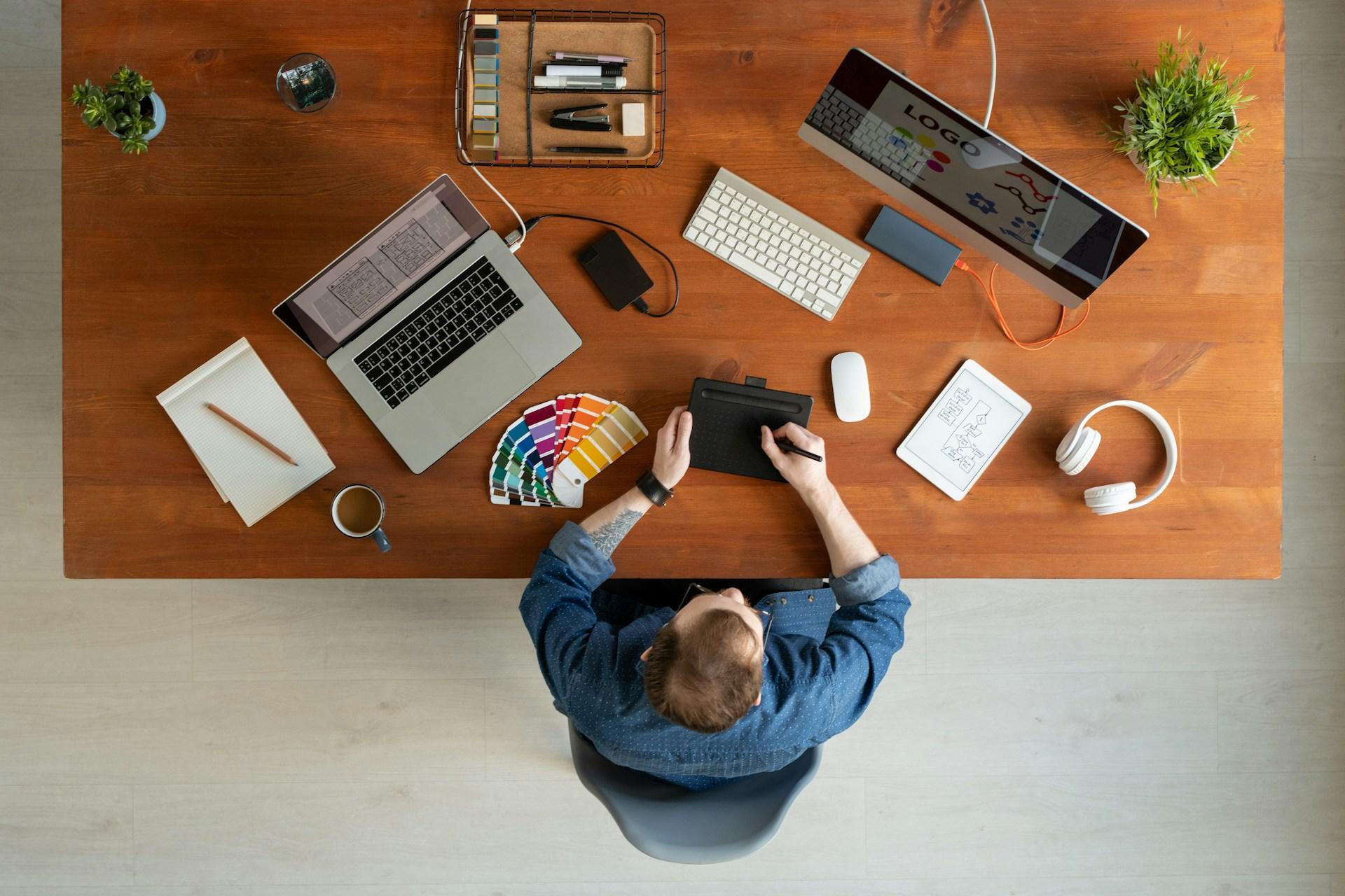 Overhead view of a designer's desk with a logo on screen, Pantone fan deck, drawing tablet and sketch pad