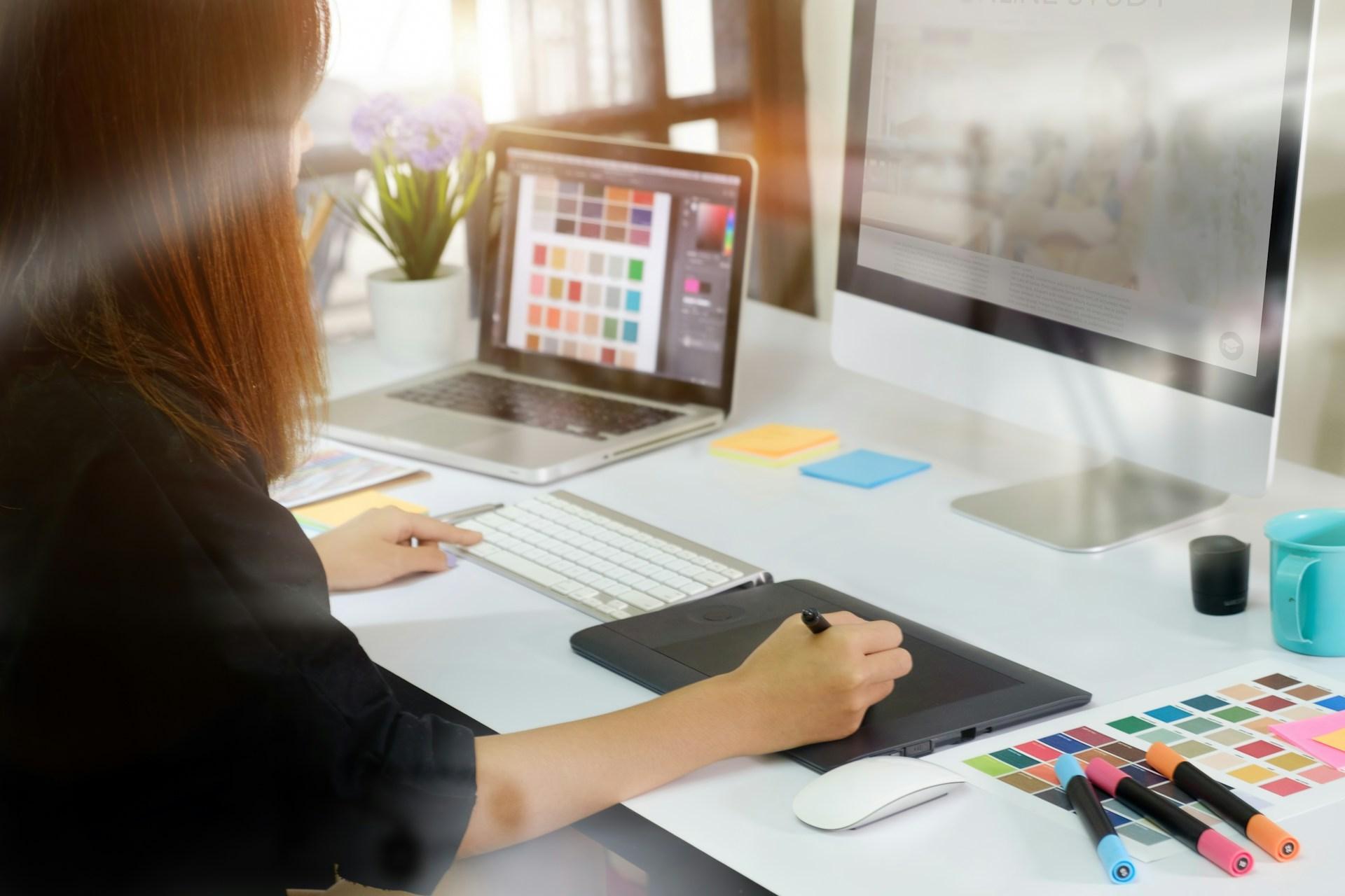Designer working on a laptop with a colour palette open, drawing tablet and printed swatch charts on the desk