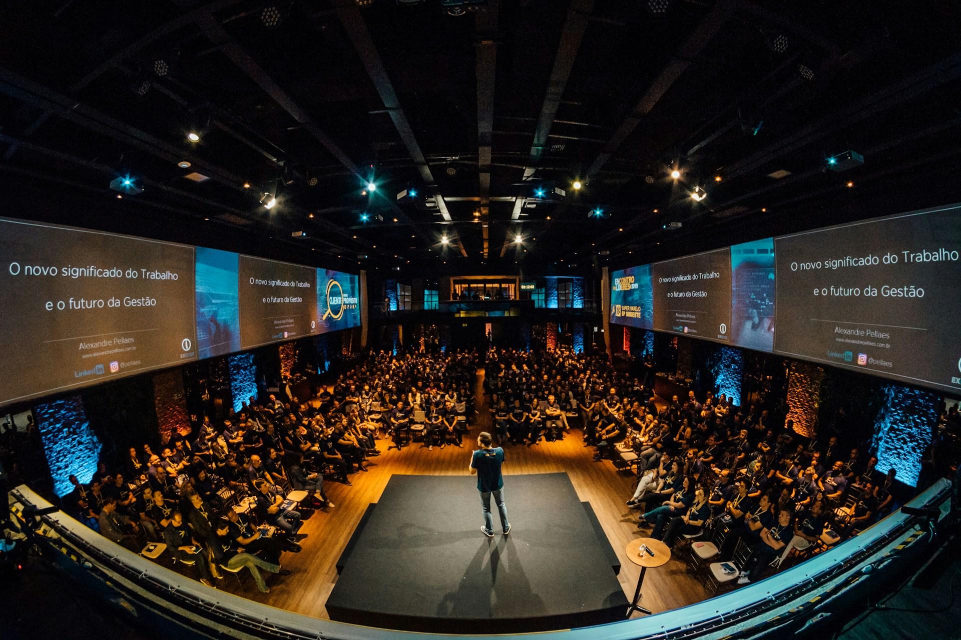 Wide-angle view of a speaker on stage at a sold-out conference with multiple screens