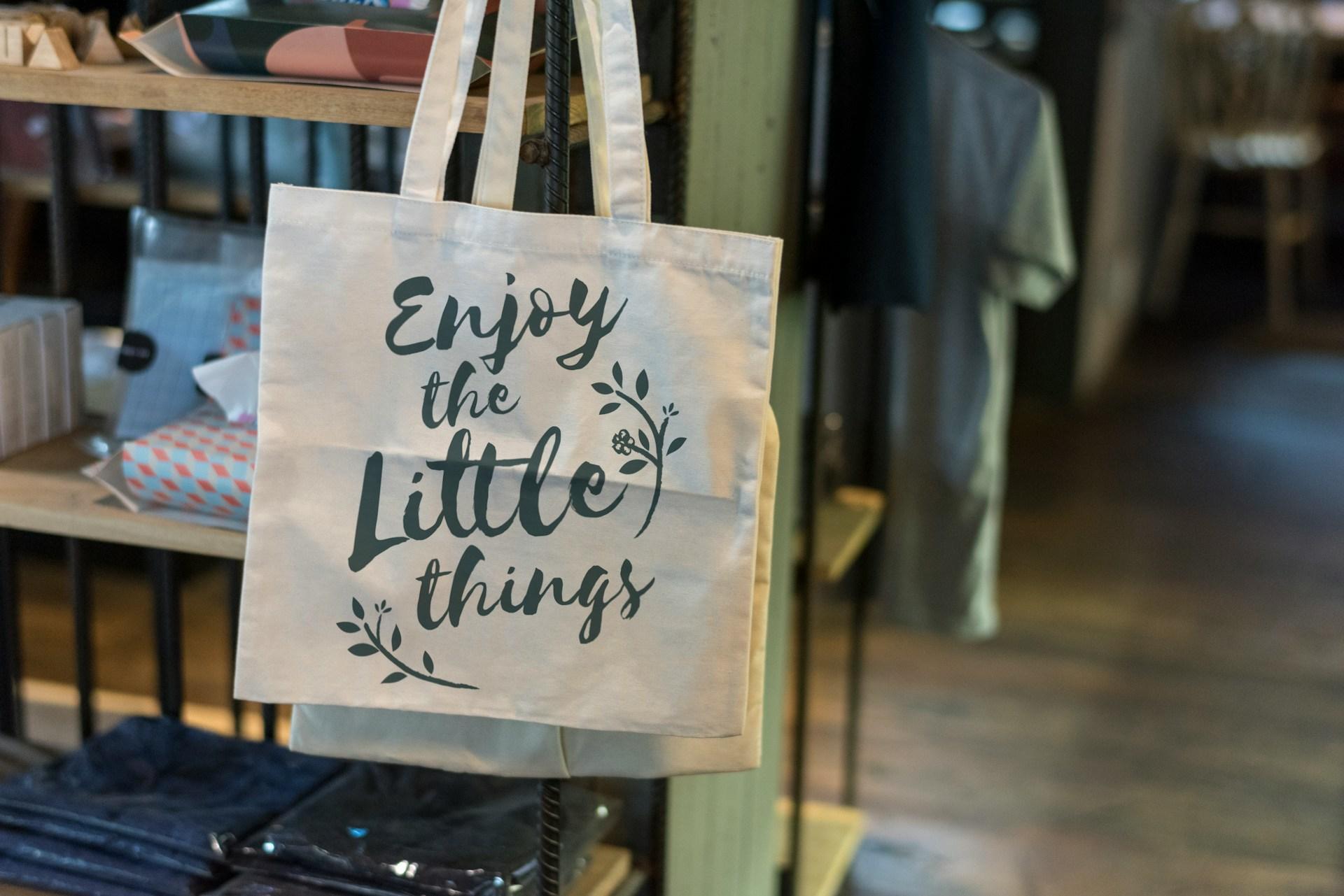 A close-up of a personalised tote bag at an expo or store, text reads "enjoy the little things"