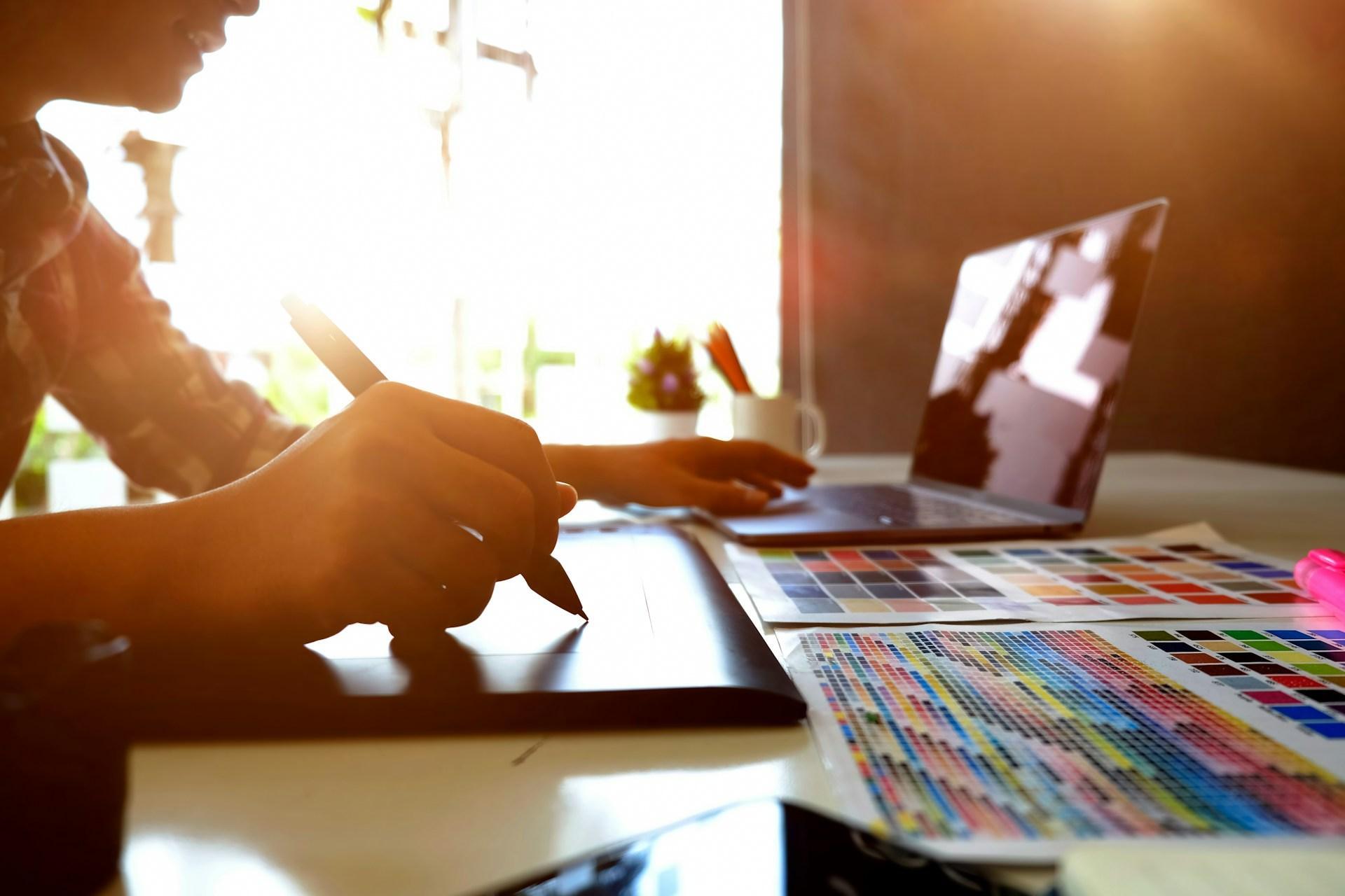 Designer redrawing artwork on a graphics tablet beside a laptop and printed colour charts