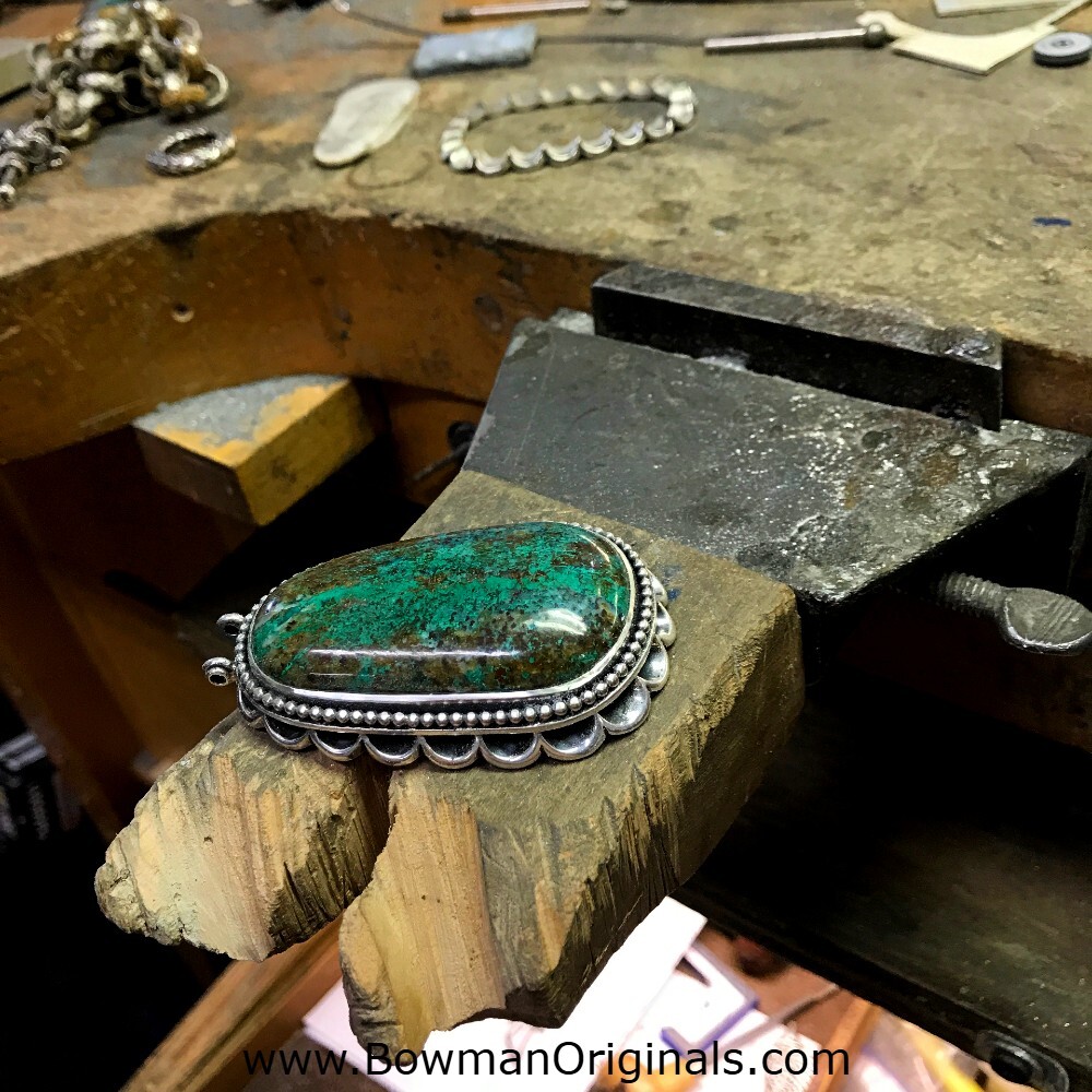 Handcut chrysocolla stone set in handmade sterling silver pendant on a workbench in the Bowman Originals workshop.