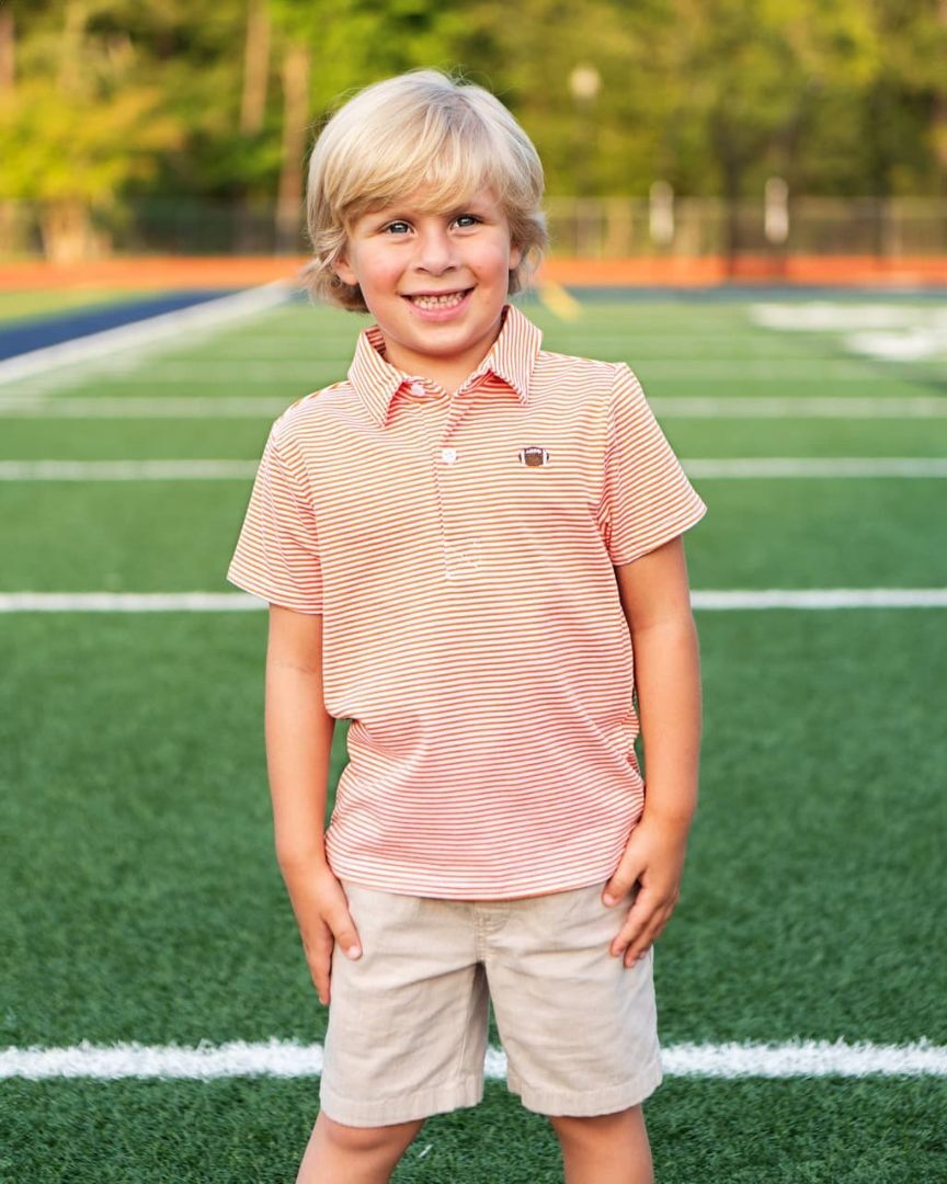 Young boy standing on a football field, wearing a light brown shirt with white stripes, ready for a tailgate or game day.