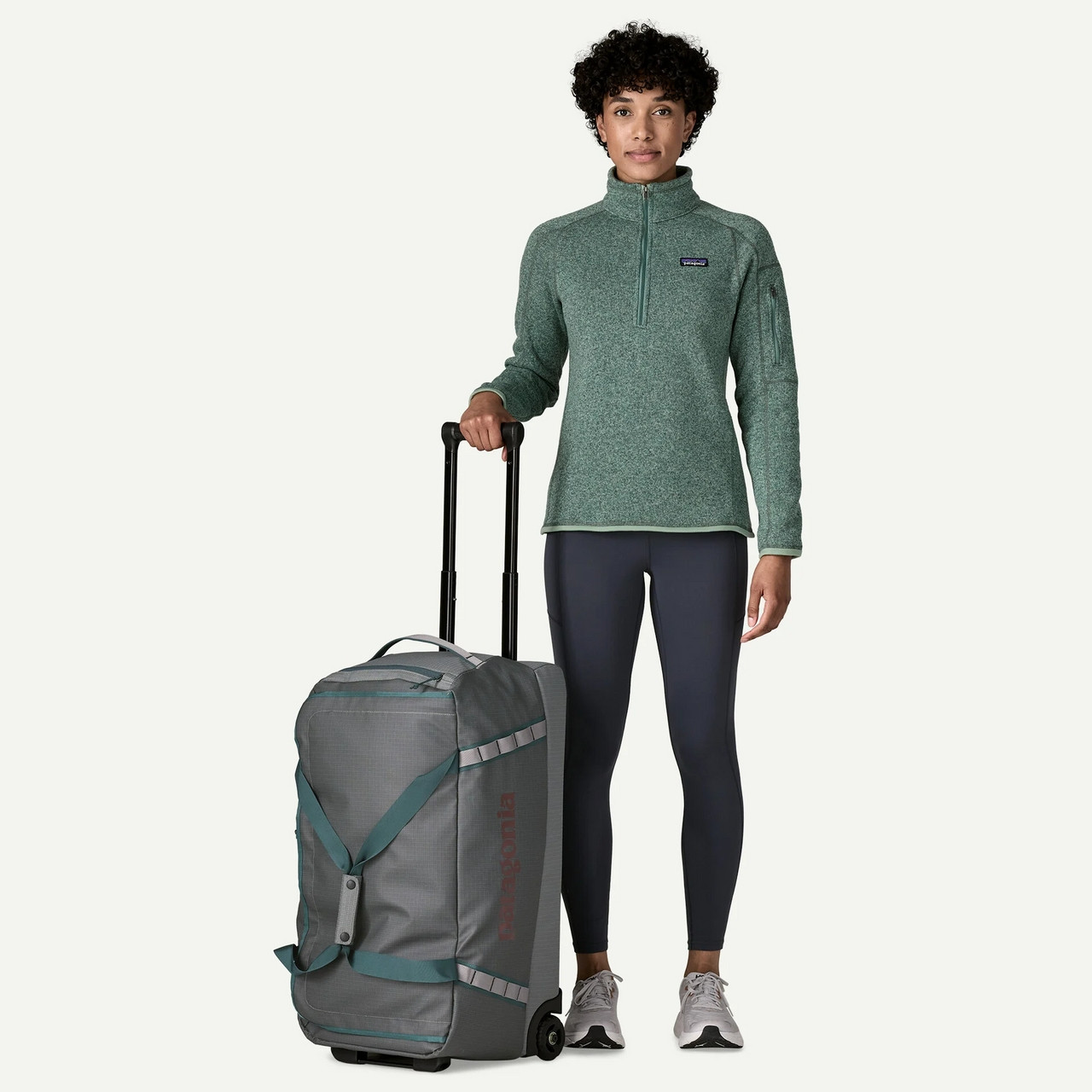 Patagonia Black Hole Wheeled Duffel 70L, a woman with short curly hair in athletic clothes standing with a gray rolling duffel bag.