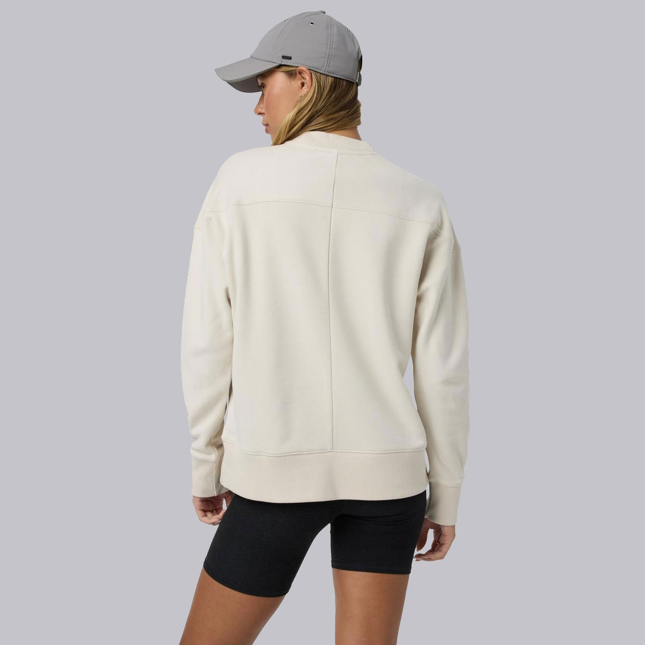 Vuori Women's Sedona Weekender Crew, woman wearing a comfortable cream-colored sweatshirt with a relaxed fit and long sleeves, paired with black shorts and a light gray cap.