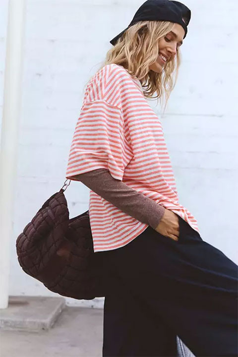 FP Movement Women's Tackle Tee - Stripe, woman wearing a red and white striped t-shirt over a brown long sleeve shirt, paired with dark pants, and a black cap, standing against a white background.
