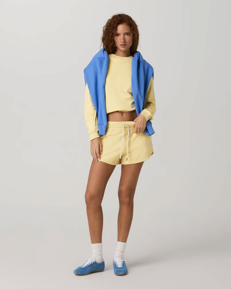 Vuori Women's Halo Modern Crew, a woman wearing a yellow cropped sweatshirt and matching yellow shorts with a blue hoodie draped over her shoulders, athletic sneakers, and white socks.