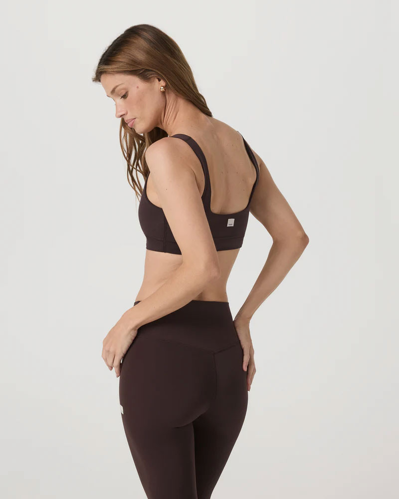 Vuori Women's AllTheForm Bra, a black athletic bra with wide straps and a supportive fit worn by a woman with long brown hair against a plain light gray background.