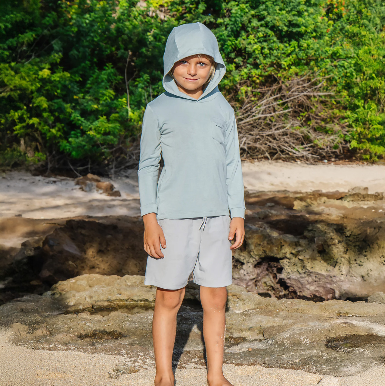 Free Fly Apparel Free Fly Toddler Breeze Short, a young boy wearing a light gray long sleeve hooded shirt and matching shorts standing on a sandy and rocky beach with green foliage in the background.