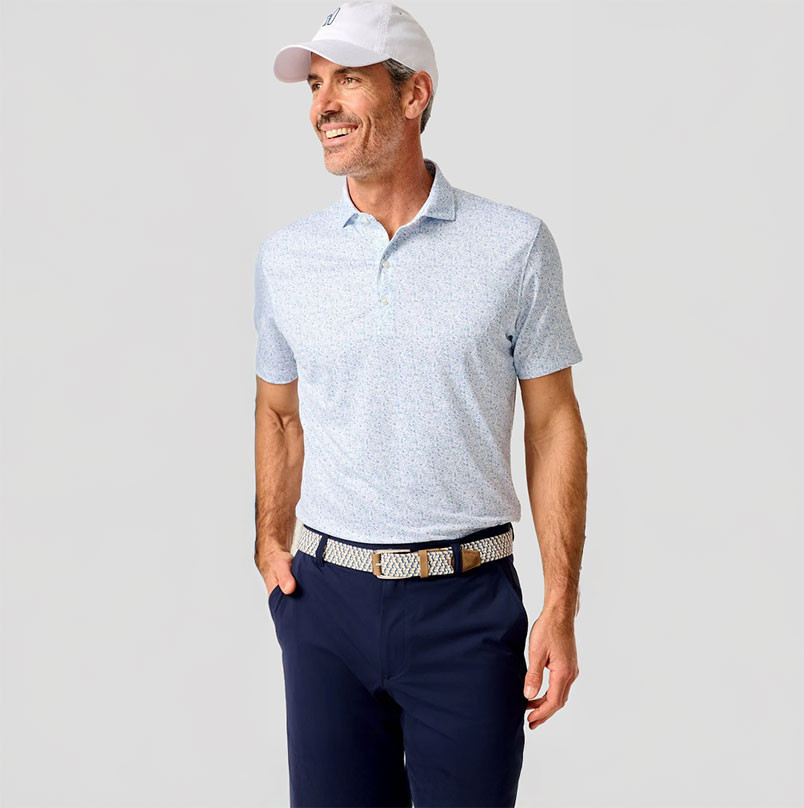 Johnnie-O Men's Performance Jersey Polo - Boggings, a man wearing a light-colored polo shirt with a subtle pattern, navy pants, a white cap, and a woven belt, standing against a plain background.