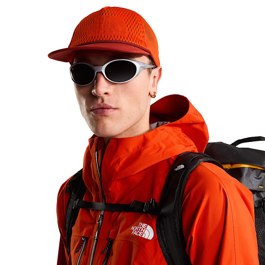 The North Face Runner Trucker, a man wearing an orange mesh-back trucker cap with a flat brim, white sporty sunglasses, and an orange waterproof jacket with a backpack, highlighting outdoor adventure gear.