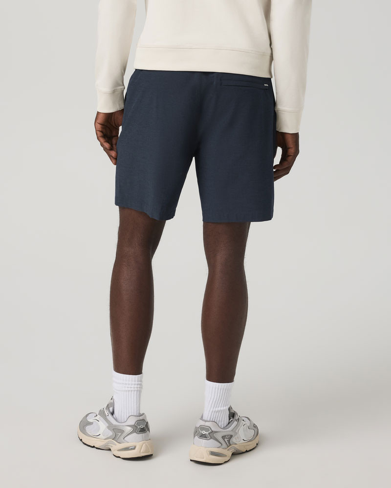 Vuori Men's Aim Elastic Waist Short - 6" Inseam, rear view of a person wearing navy blue shorts with a rear pocket, paired with white sneakers and white socks.