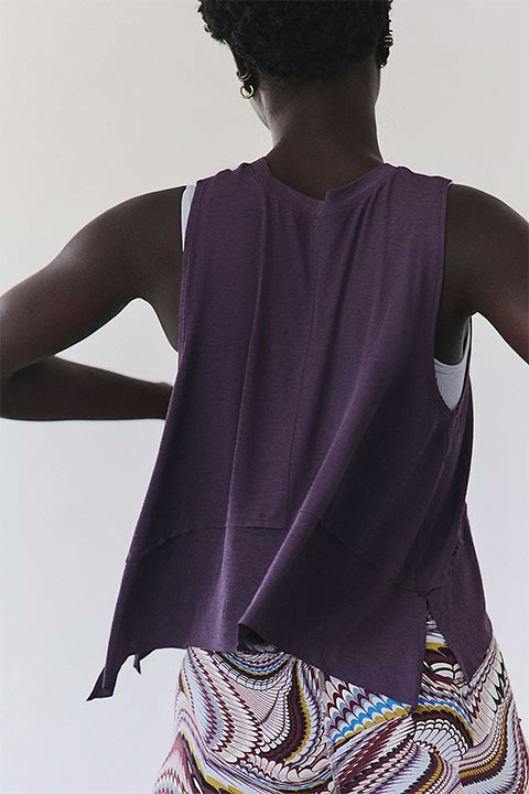 FP Movement Women's Tempo Tank, back view of a woman wearing a purple sleeveless athletic tank top with a white sports bra underneath, paired with colorful patterned workout shorts.