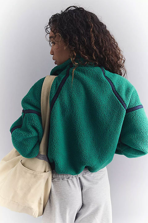 FP Movement Women's Mountain High 1/2 Zip Pullover, a woman wearing a teal fleece pullover with navy accents and a beige tote bag.
