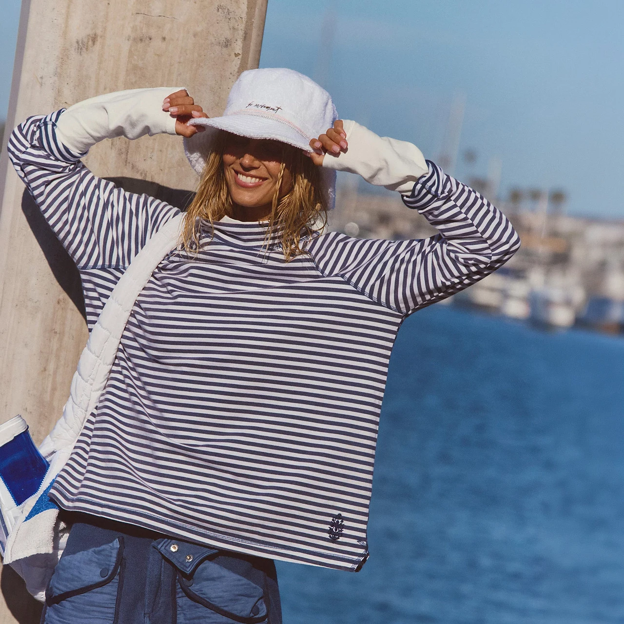 FP Movement Women's Restoration Yarn Dye Stripe Layer, a stylish women's striped layered top in navy and white with a small anchor logo at the hem, worn by a smiling woman at a seaside location.