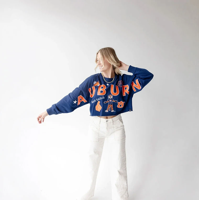 Gameday Social Apparel Gameday Social Auburn Patched Up Cropped LS Tee, model wearing a navy cropped long-sleeve Auburn sweatshirt with orange and white graphics and patches, styled with white jeans.