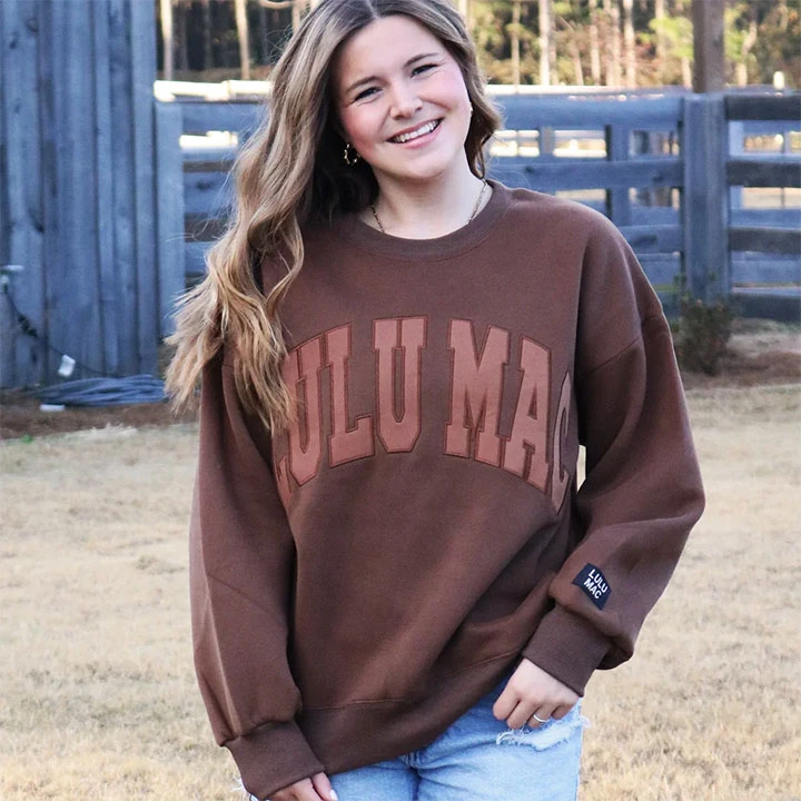 Lulu Mac Women's Mono Sweatshirt, a young woman wearing a brown sweatshirt with large 'LULU MAC' lettering and a small brand patch on the sleeve, standing outdoors on grass with a wooden fence in the background.