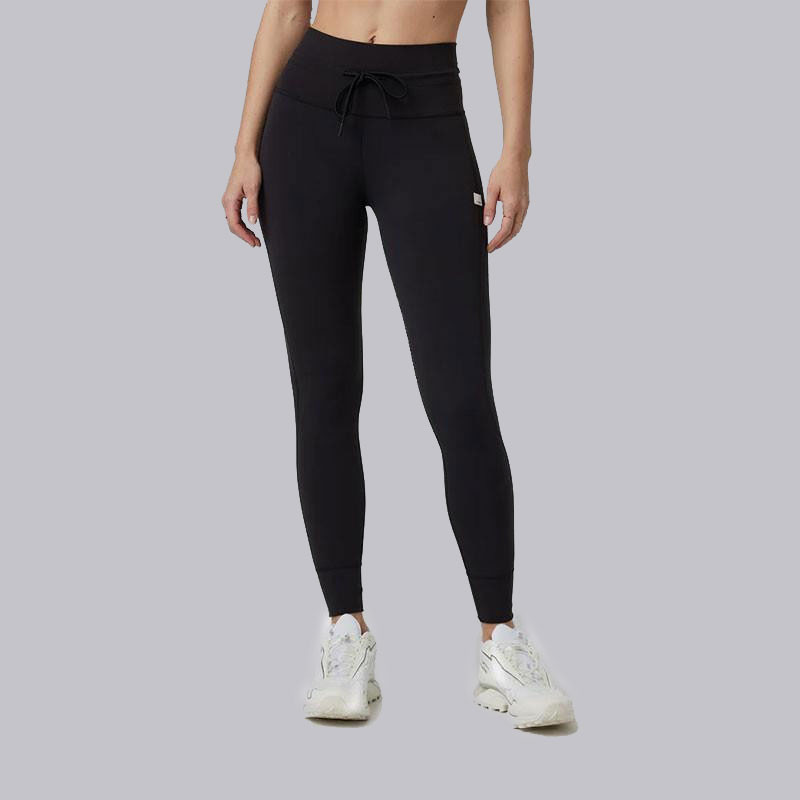 Vuori Women's AllTheFeels Legging, black high-waisted athletic leggings with a drawstring waist and slim fit, paired with white sneakers.