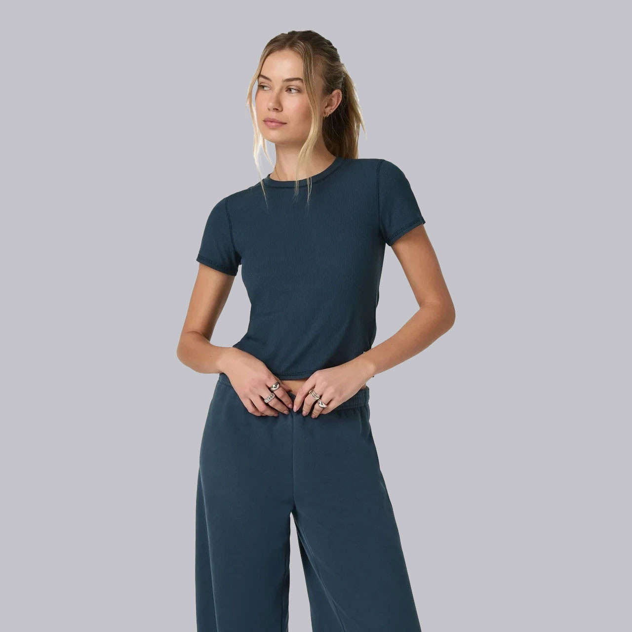 Vuori Women's Pose Fitted Tee, a woman wearing a navy blue fitted T-shirt with short sleeves and a round neckline, paired with matching wide-leg pants, standing against a light gray background.