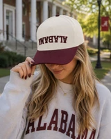 Alabama Outdoors Captuer Upside Down Alabama Logo Snapback, a woman wearing a white and maroon baseball cap with 'Alabama' embroidered upside down on the front, outdoors in a campus setting.