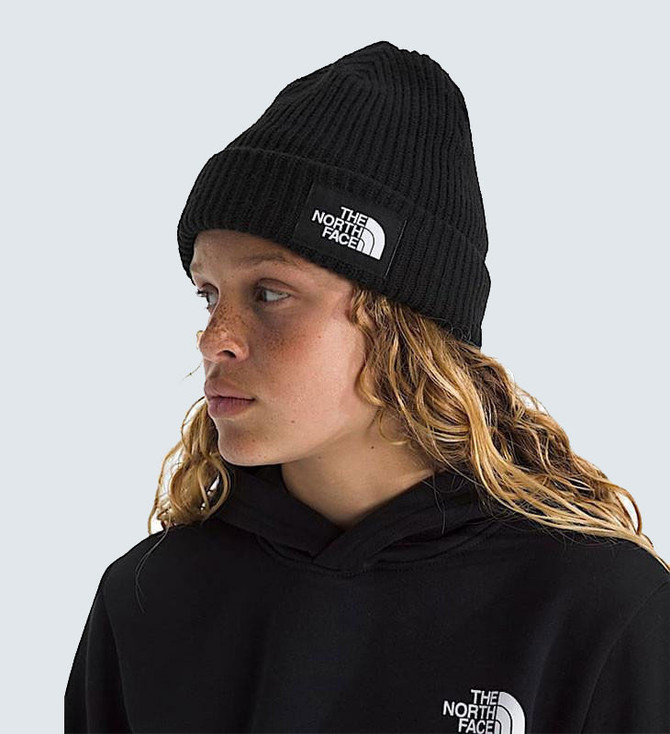 Summer Sale exclusions The North Face Kids' Salty Lined Beanie, black knitted beanie with white logo patch, worn by a woman with curly blonde hair and a black hoodie, against a light grey background.