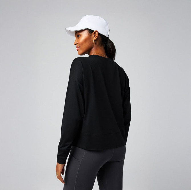 Fabletics Women's Cloud Jersey Boxy Long-Sleeve, image of a woman in a black long-sleeve top with a relaxed fit, paired with dark leggings, wearing a white cap, and posing against a plain background.