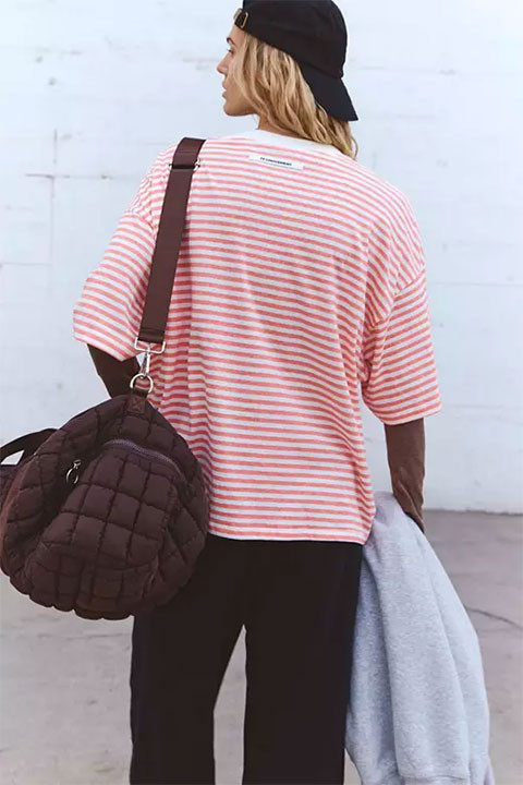 FP Movement Women's Tackle Tee - Stripe, a woman wearing a pink and white striped T-shirt, black cap, carrying a backpack and holding a gray sweatshirt.