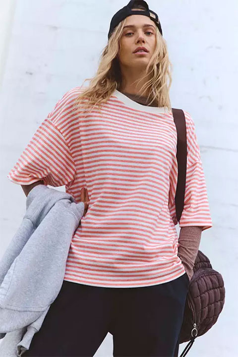FP Movement Women's Tackle Tee - Stripe, woman wearing casual striped t-shirt with pink and white horizontal stripes, black pants, and a black cap, carrying a gray jacket and a quilted bag.