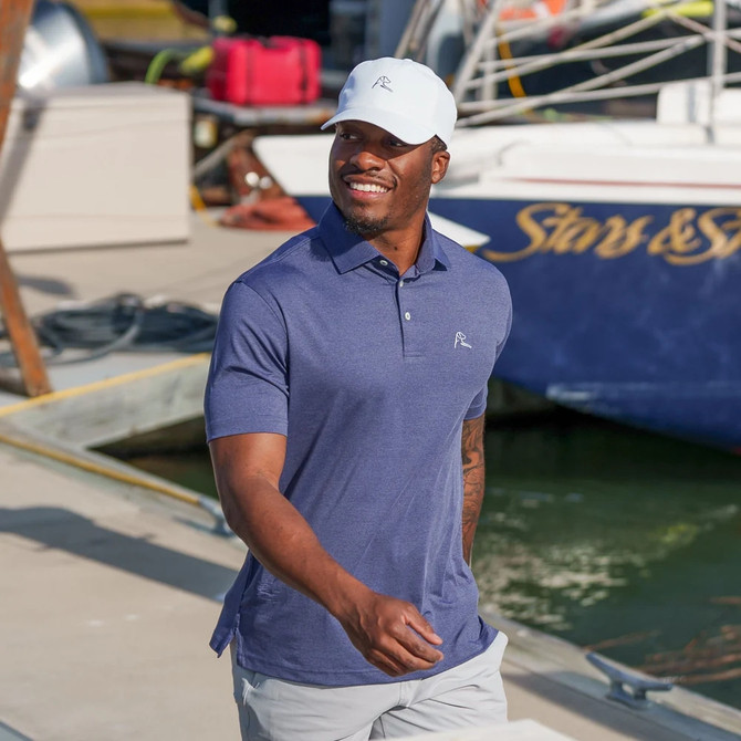 Rhoback Men's Heather Performance Polo, a man wearing a blue performance polo shirt with a small logo on the chest, smiling while walking near a yacht docked at a marina.