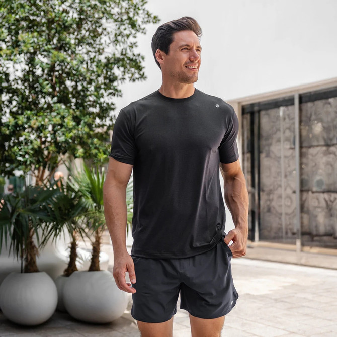Rhoback Men's Blitz Tech Tee, a man wearing a black athletic T-shirt outdoors with plants and a modern building in the background.