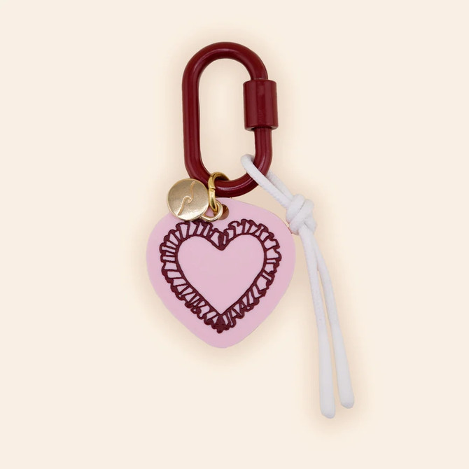 Freshwater Curated Goods Ruffled Heart Bag Charm, pink heart-shaped accessory with ruffled outline, attached to a maroon clasp with a small gold charm and white cord for decoration.