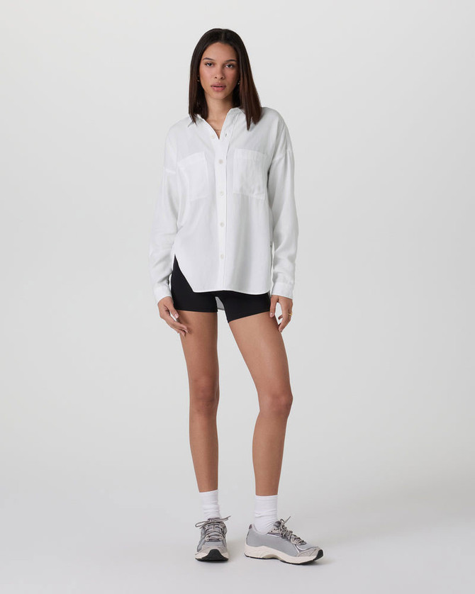 Vuori Women's Trestles Twill Button Up, a woman wearing a white twill button-up shirt paired with black shorts and athletic sneakers, standing against a plain background.